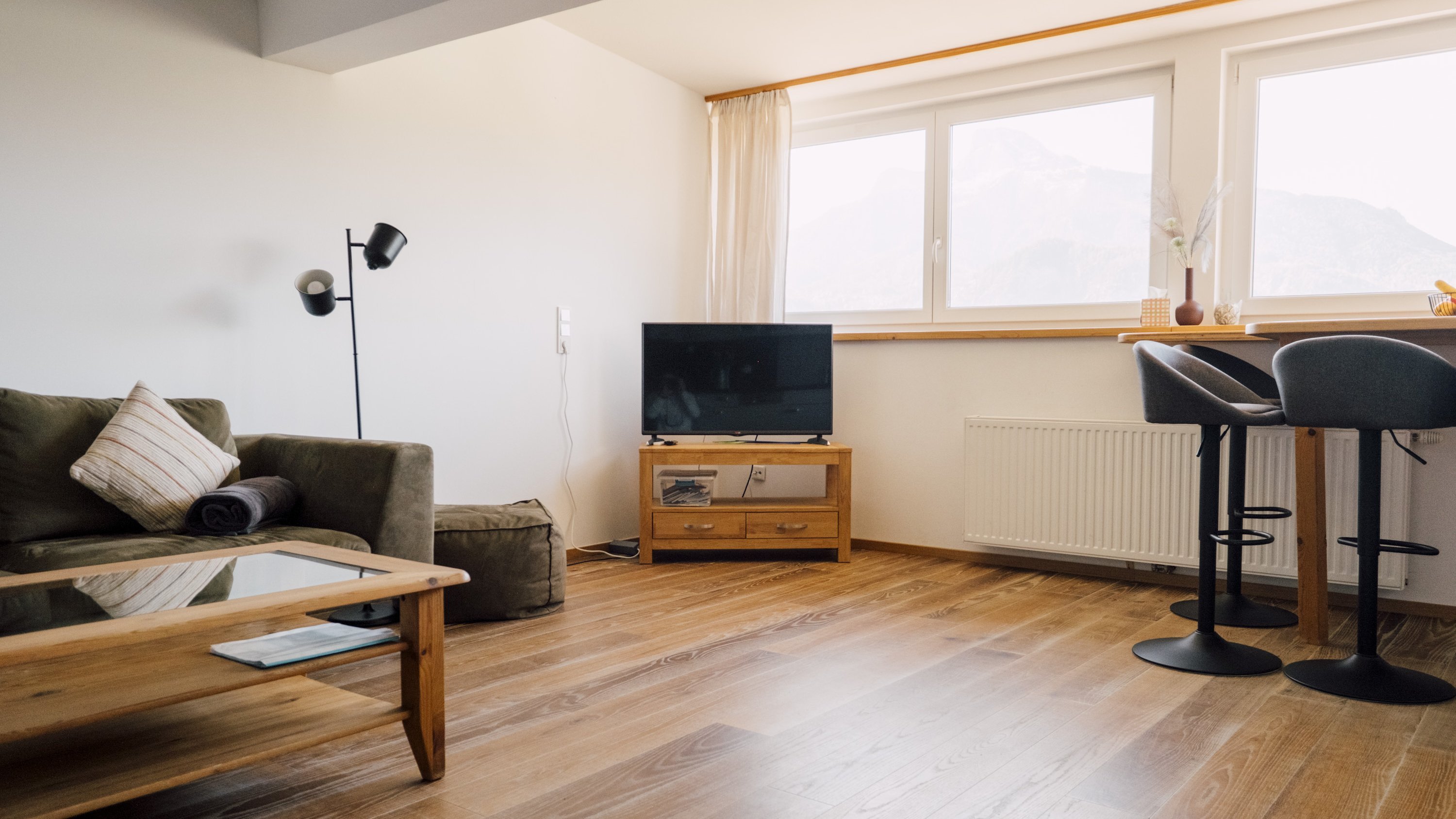 Heller, moderner Wohnraum mit Sofa, Fernseher und Essbereich mit Barhockern sowie großen Fenstern.