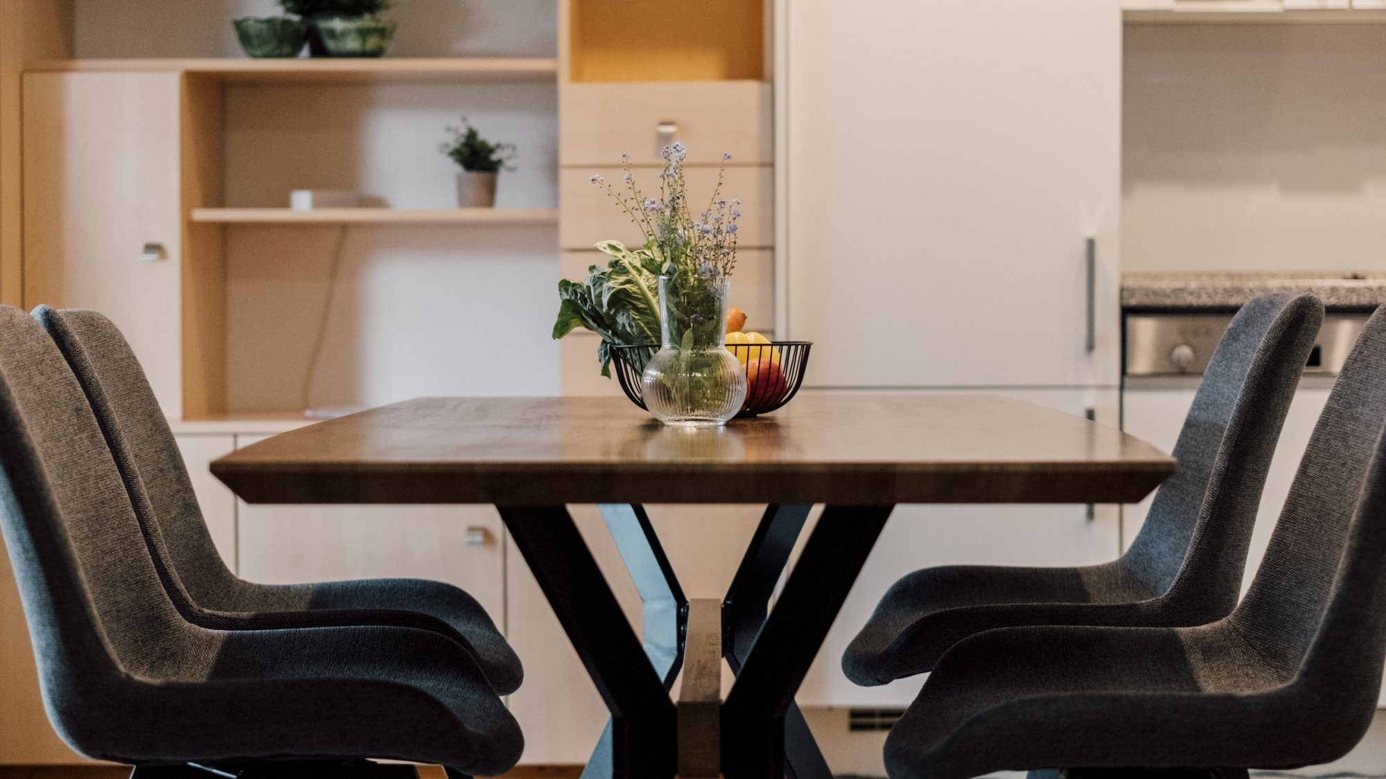 Esszimmer mit Holztisch und grauen Stühlen vor hellen Küchenschränken und Geräten