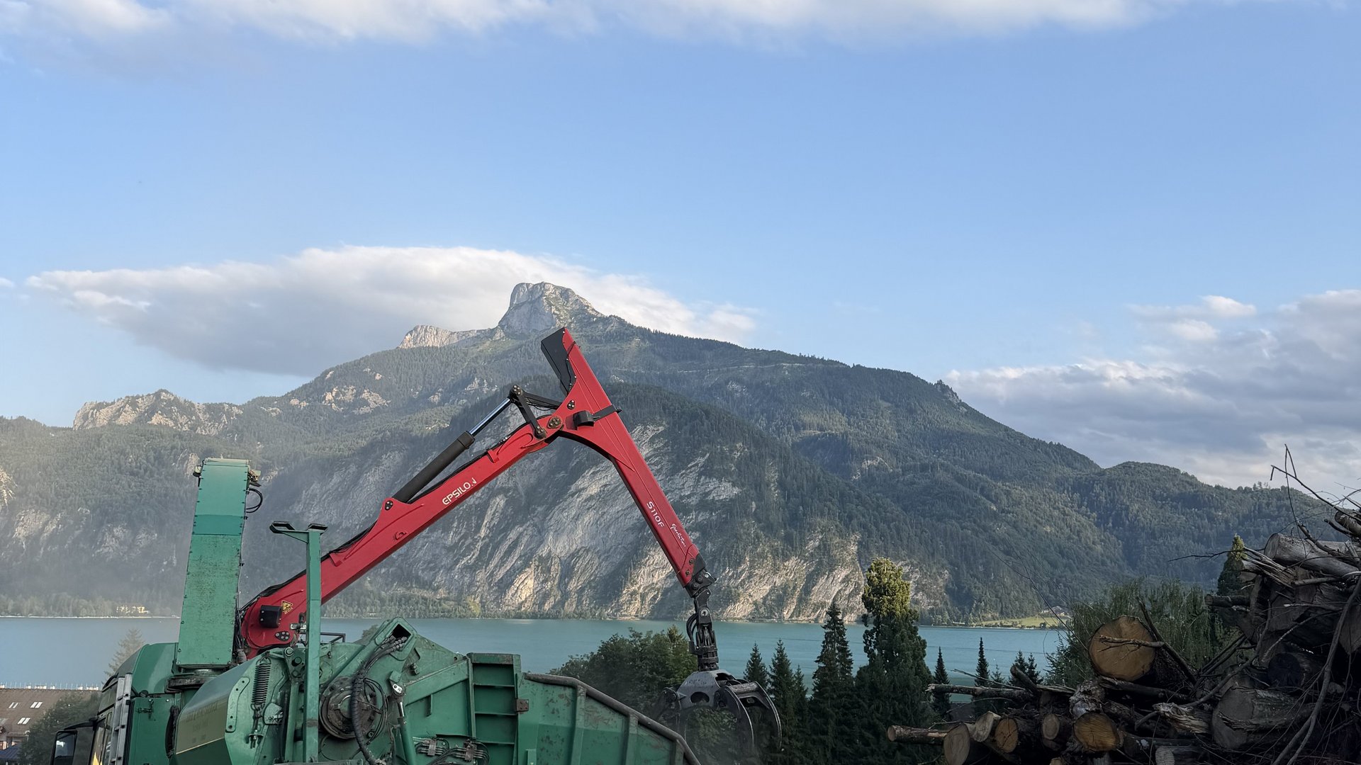 Forstwirtschaft im Gebirge Ein Holzerntefahrzeug mit Kran in einer Berglandschaft vor einem See, umgeben von gestapelten Stämmen.
