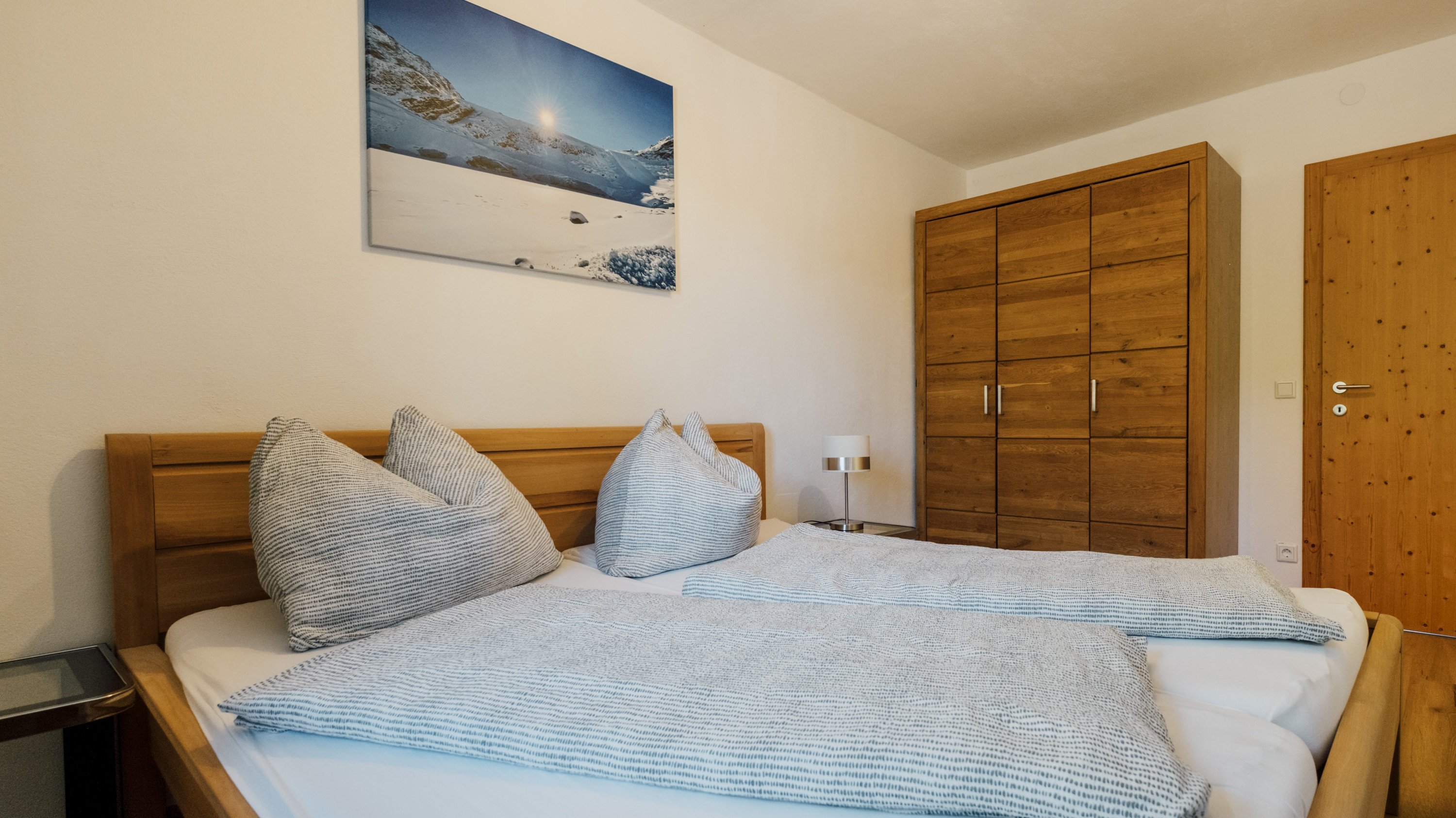 Helles Schlafzimmer mit einem Holzbett, dekorativen Kissen und Kunstwerk einer Berglandschaft an der Wand.