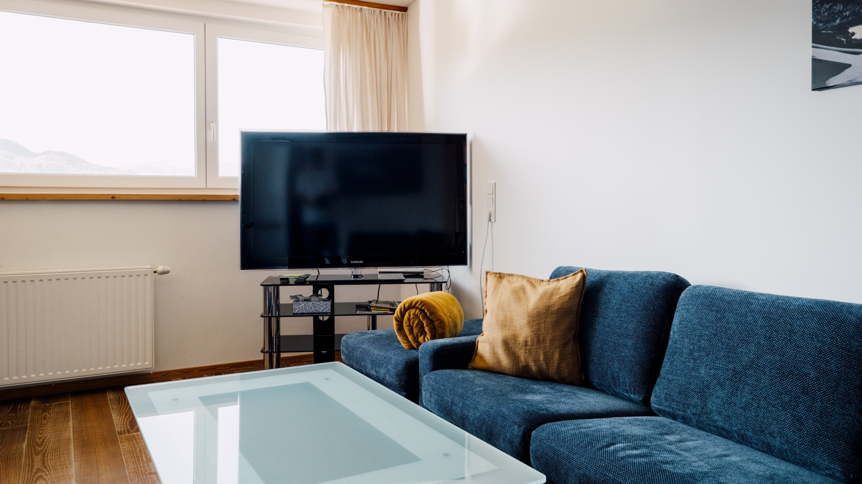 Ein modernes Wohnzimmer mit einem blauen Sofa, Glastisch und Fernseher auf einem Stand.