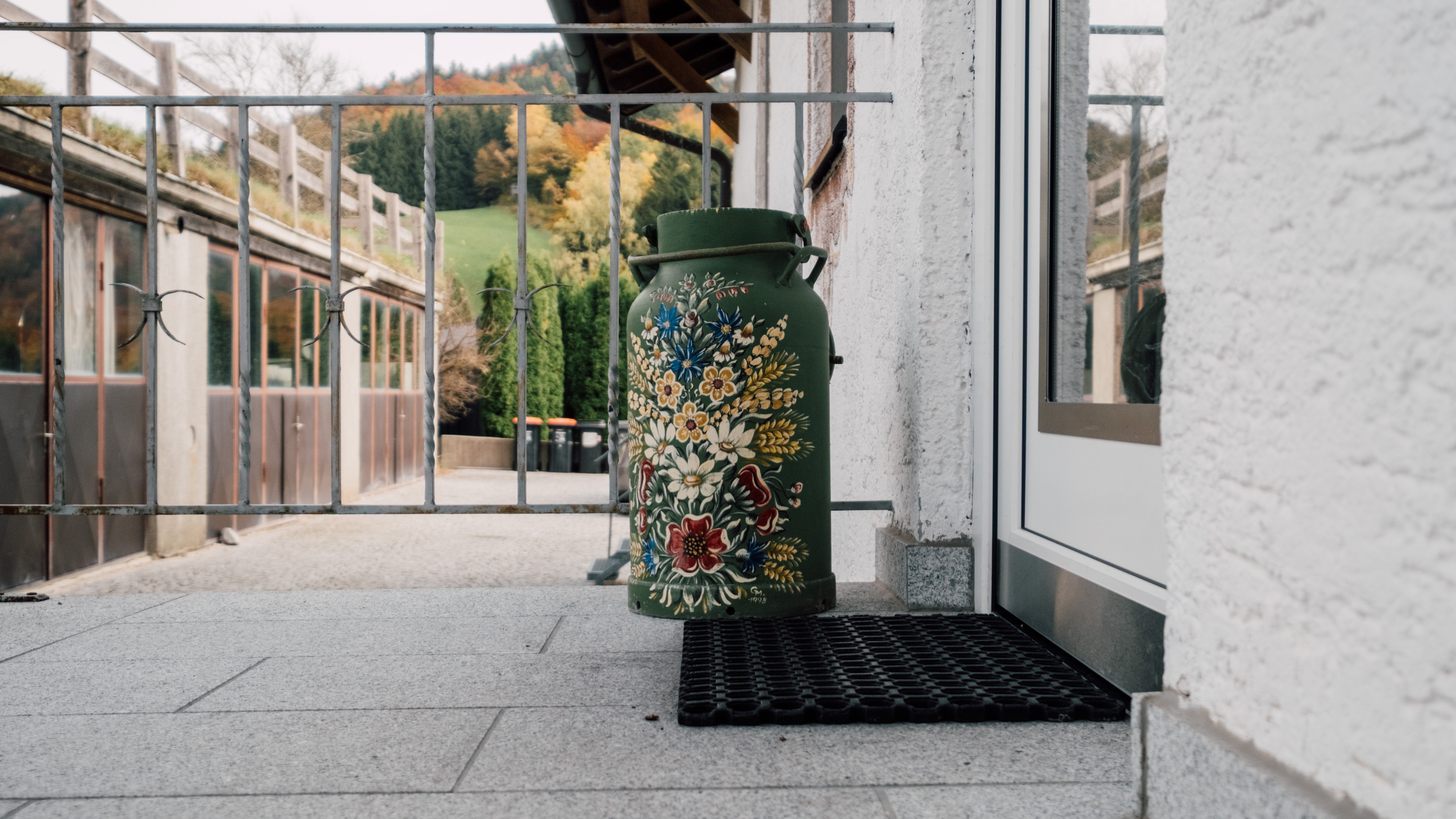 Dekorative Milchkann in rustikalem Design an einem Außeneingang mit Matten und Zaun im Hintergrund.