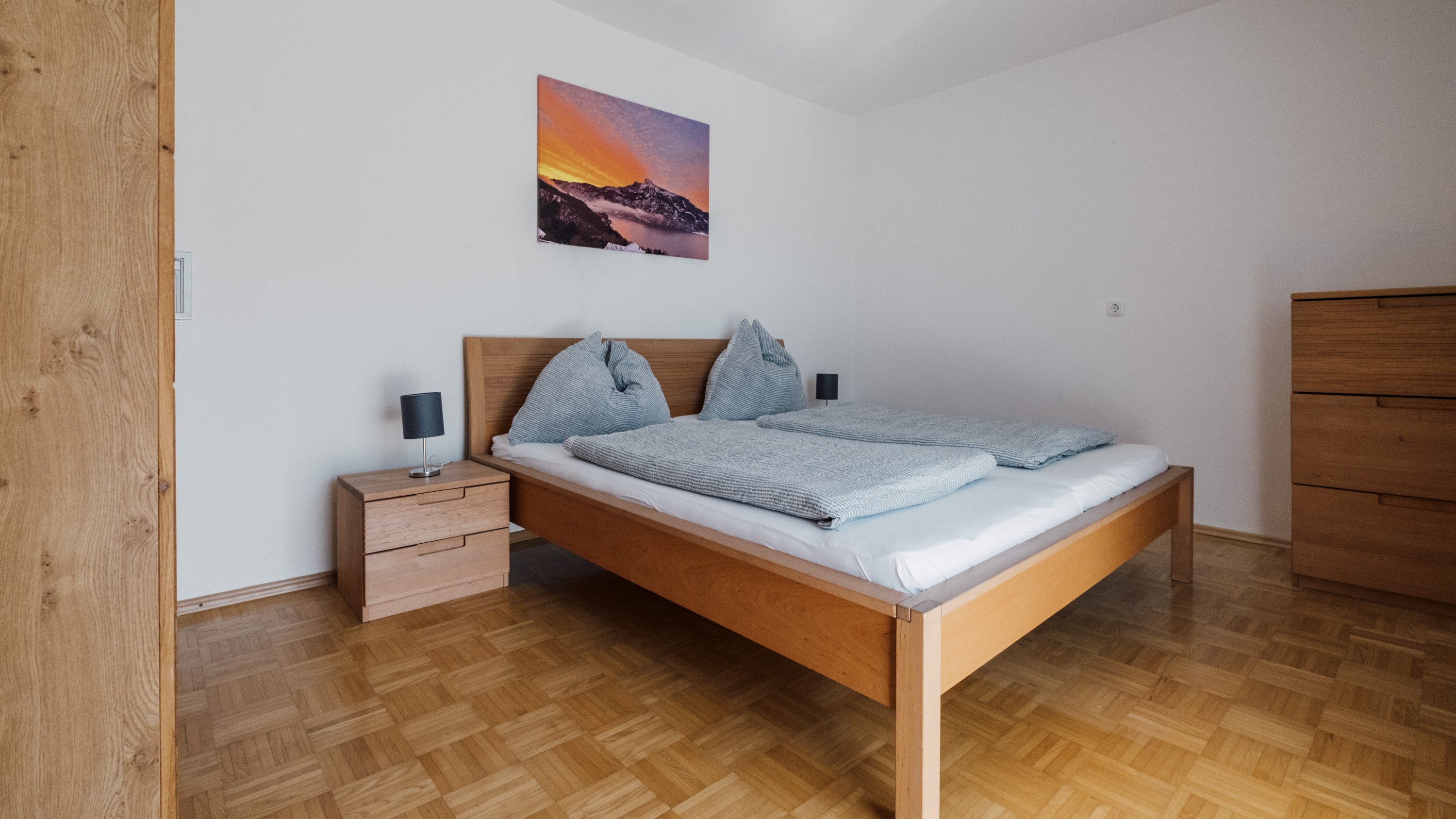 Modernes Schlafzimmer mit Holzbett, Kissen, Tagesdecke und Landschaftsgemälde an der Wand