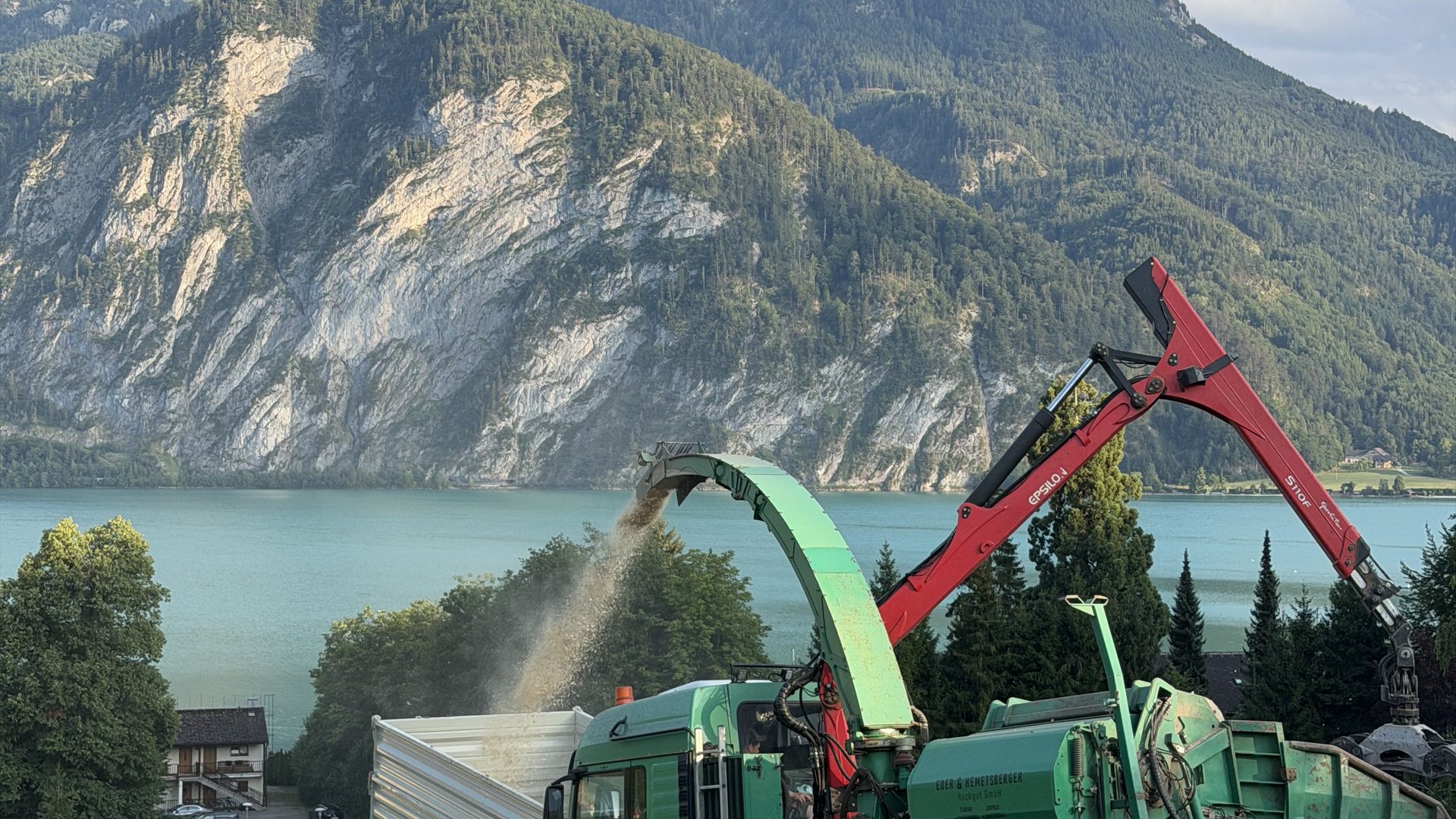 Hackmaschine mit rotem Kranarm bläst frische Hackschnitzel in einen Container, im Hintergrund der Mondsee und eine Felswand