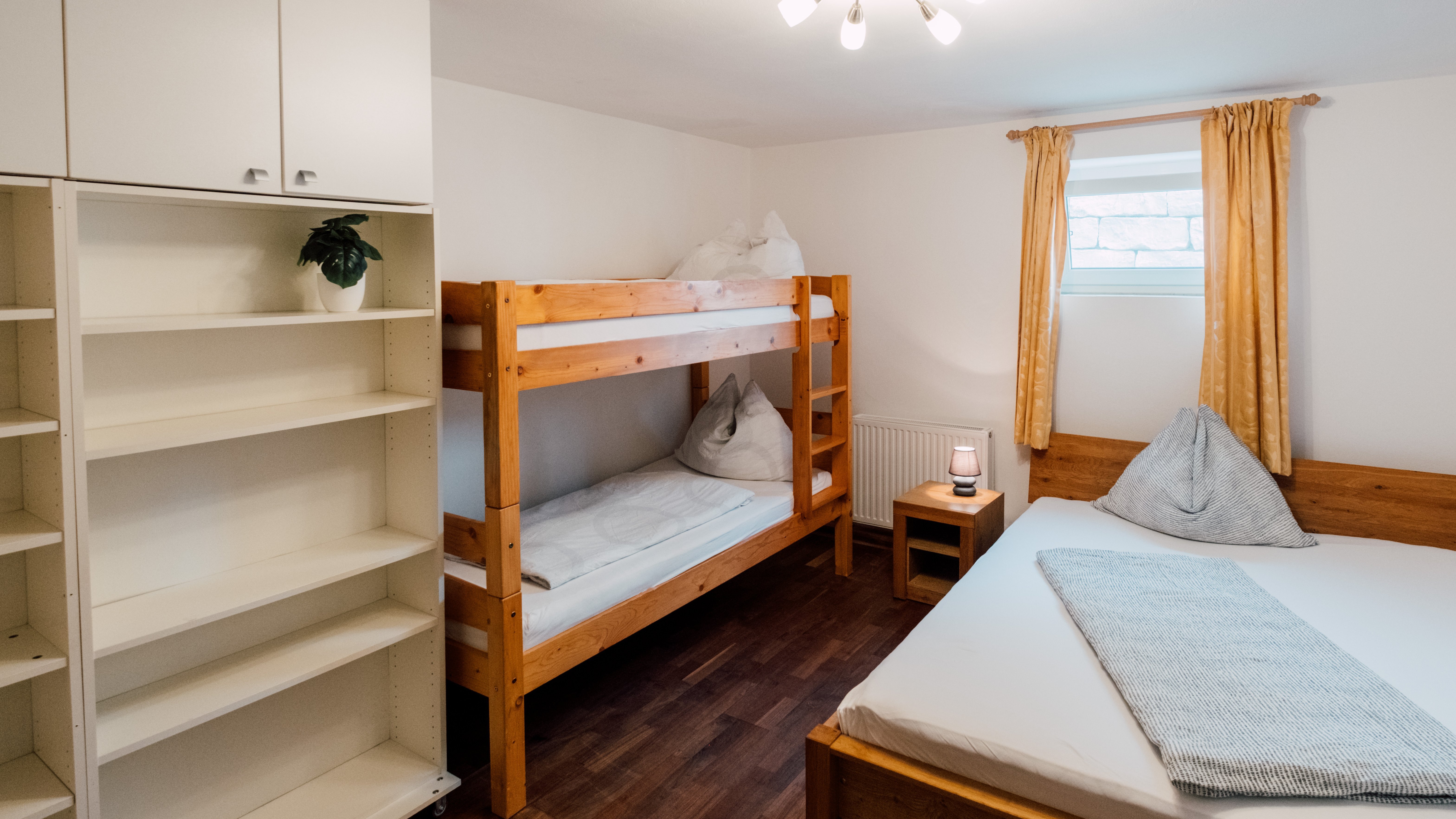 Ein Schlafzimmer mit Etagenbett und Doppelbett sowie einem Regal im Raum, beleuchtet durch Tageslicht.