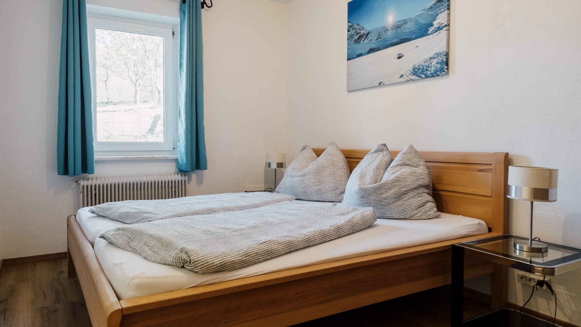 Helles Schlafzimmer mit Holzbett und grauen Kissen, Fenster mit blauen Vorhängen.