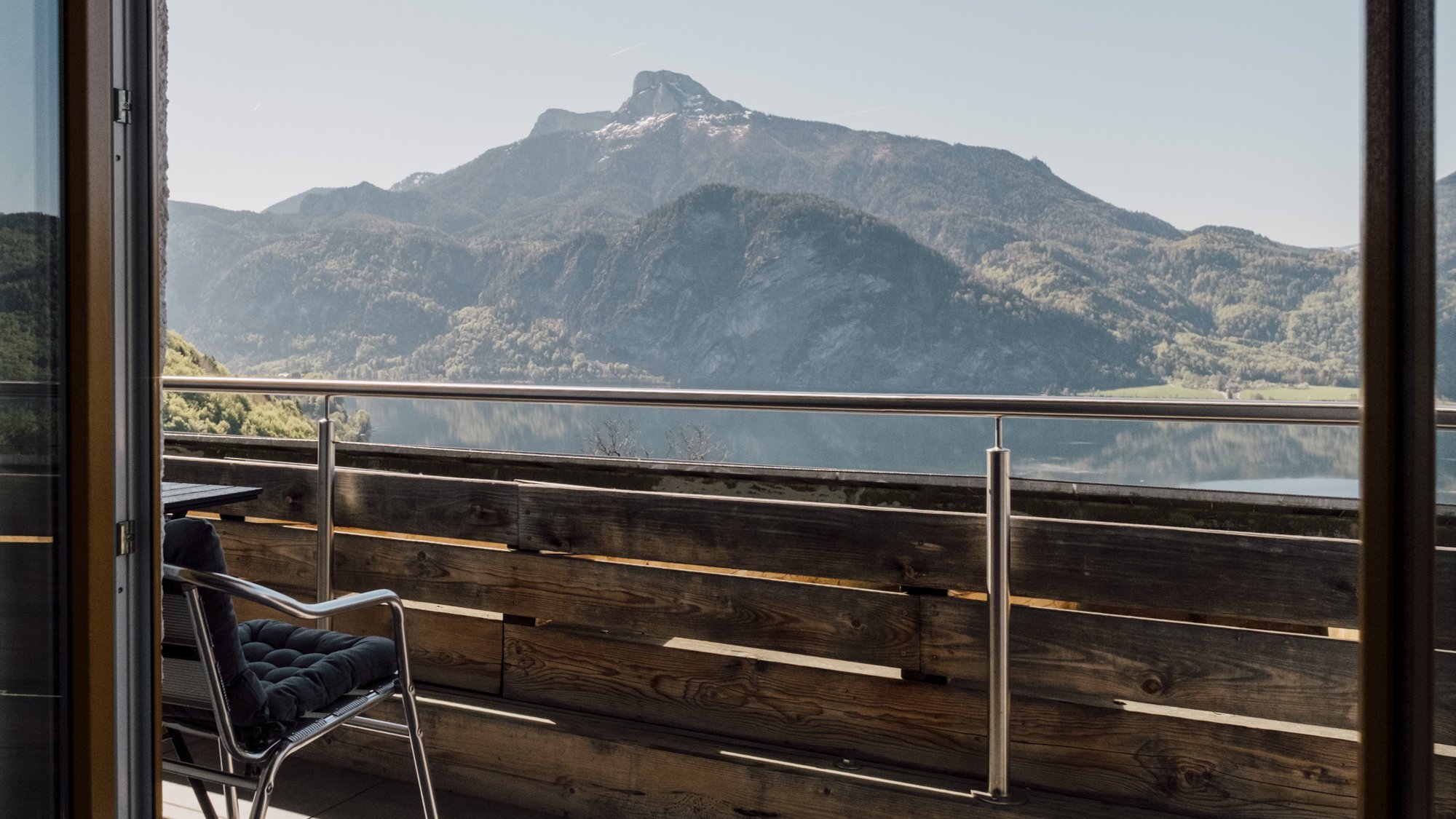 Atemberaubender Blick auf die Berge Blick durch eine große Glastür auf einen Balkon mit einem Stuhl und Berglandschaft im Hintergrund
