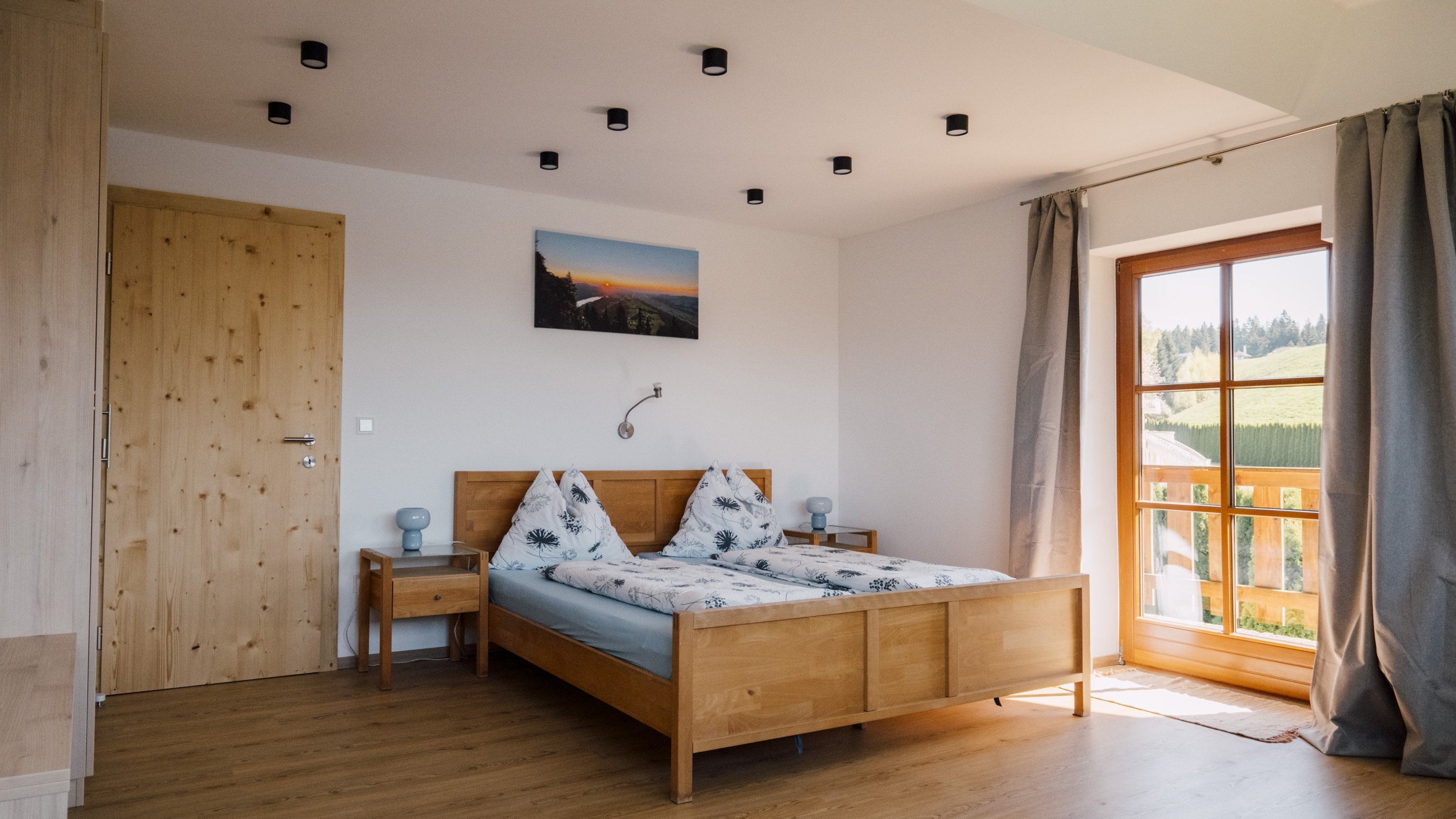 Helles Schlafzimmer mit einem Holzbett, Dekokissen und großer Fensterfront mit Ausblick