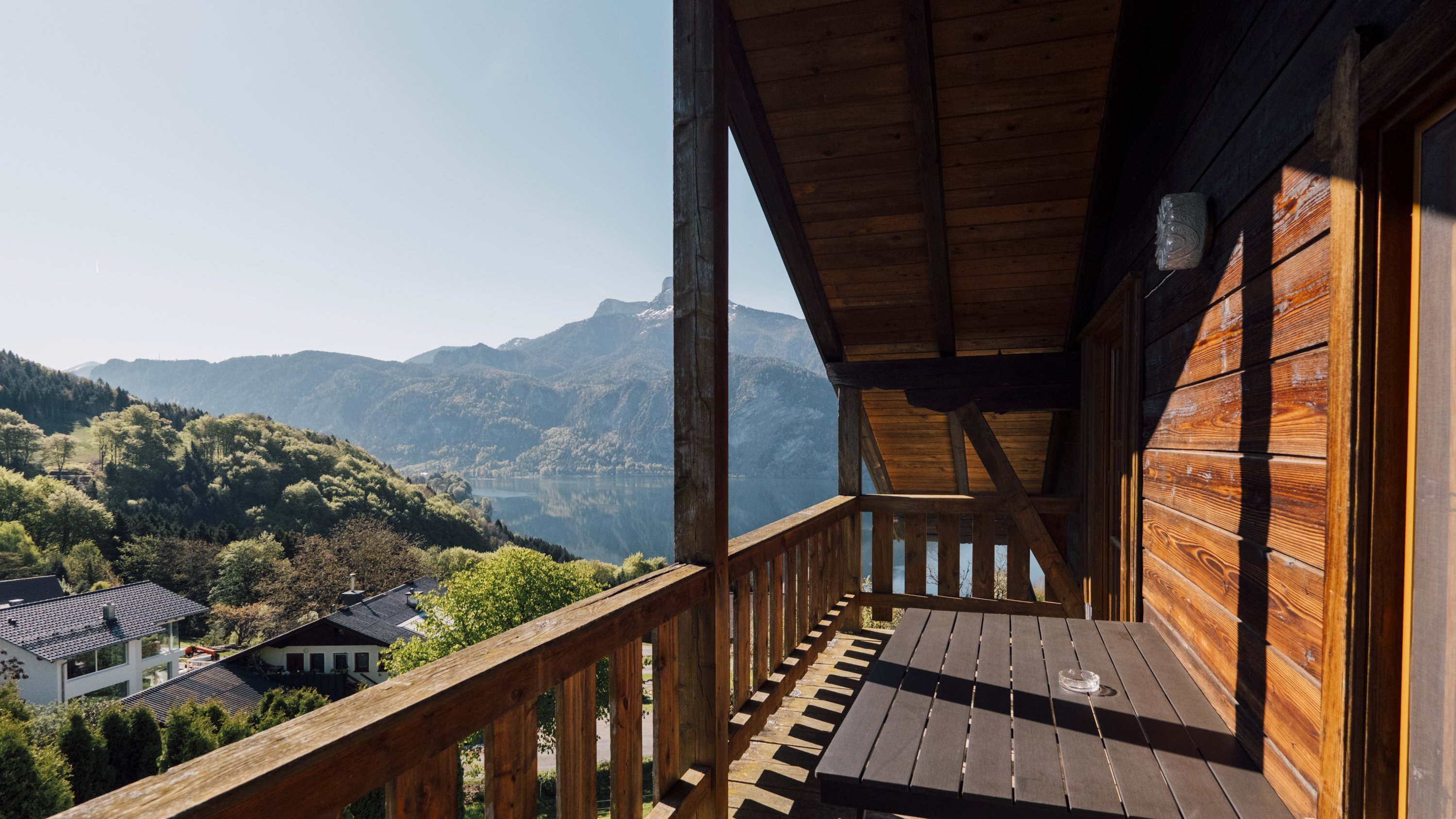 Holzbalkon mit Blick auf eine Berglandschaft und einen See im Vordergrund unter blauem Himmel.