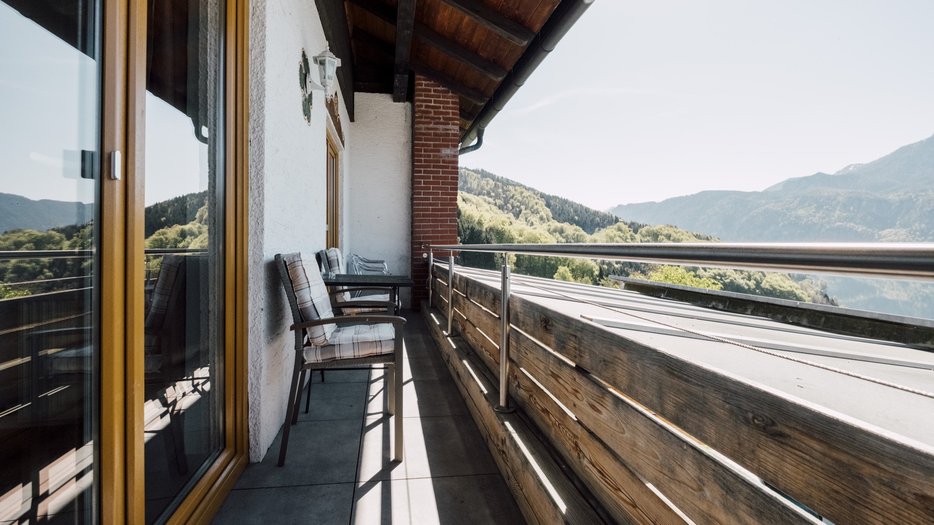 Ein Balkon mit Holzgeländer und Sitzgelegenheiten überblickt eine Berglandschaft unter klarem Himmel.