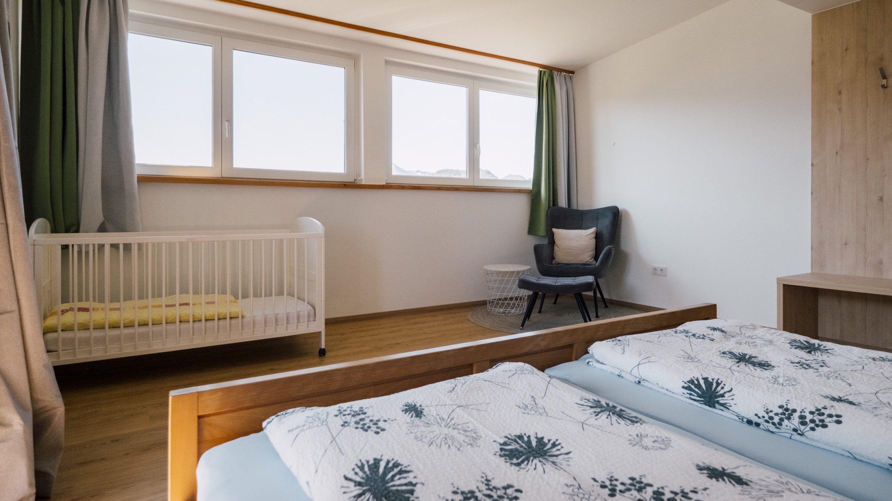 Ein modern gestaltetes Schlafzimmer mit einem Babybett und einem Doppelbett, große Fenster für viel Tageslicht.