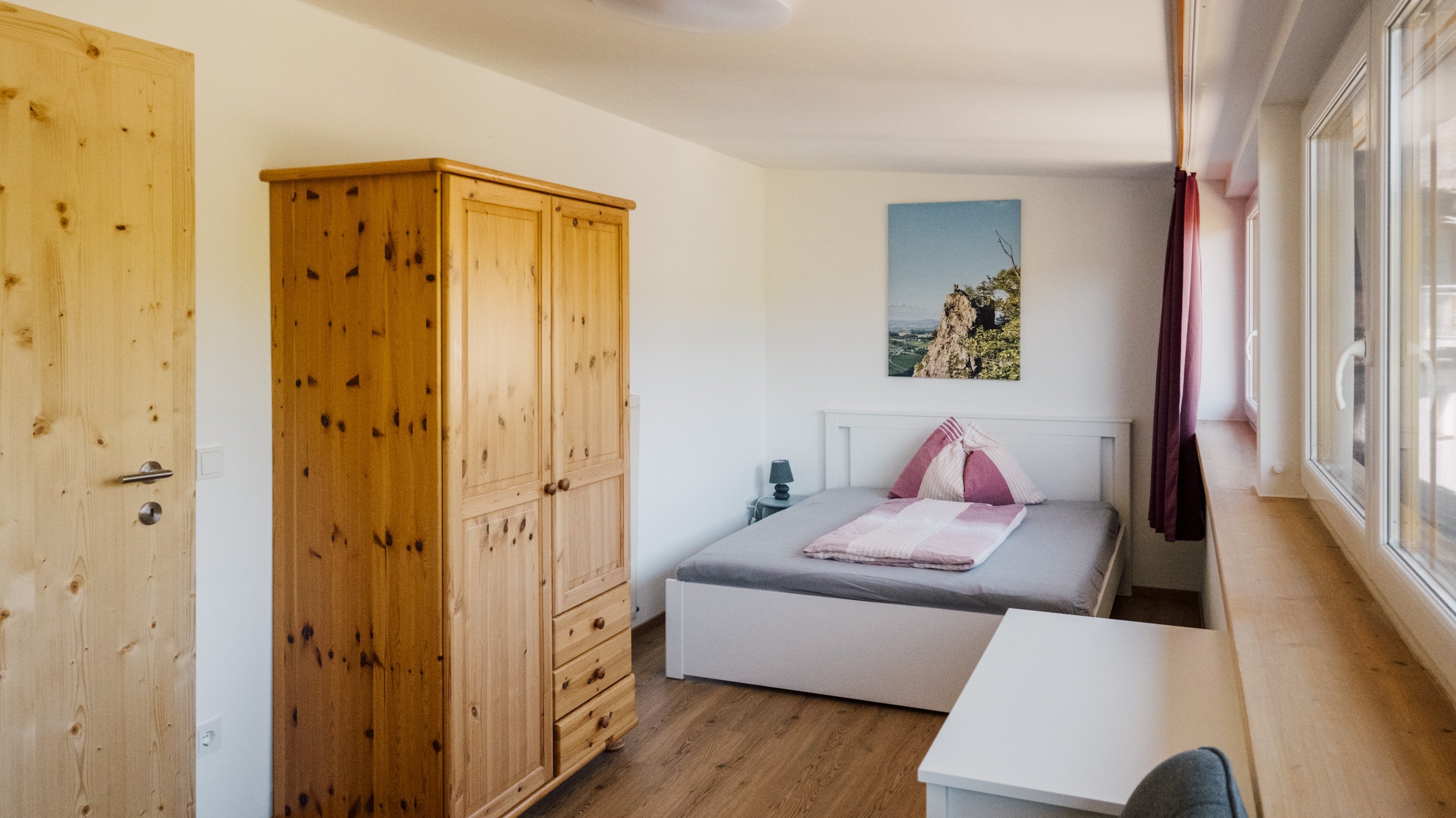 Ein helles Schlafzimmer mit einem Bett, einem Holzschrank und einem Schreibtisch sowie einem Landschaftsbild an der Wand.