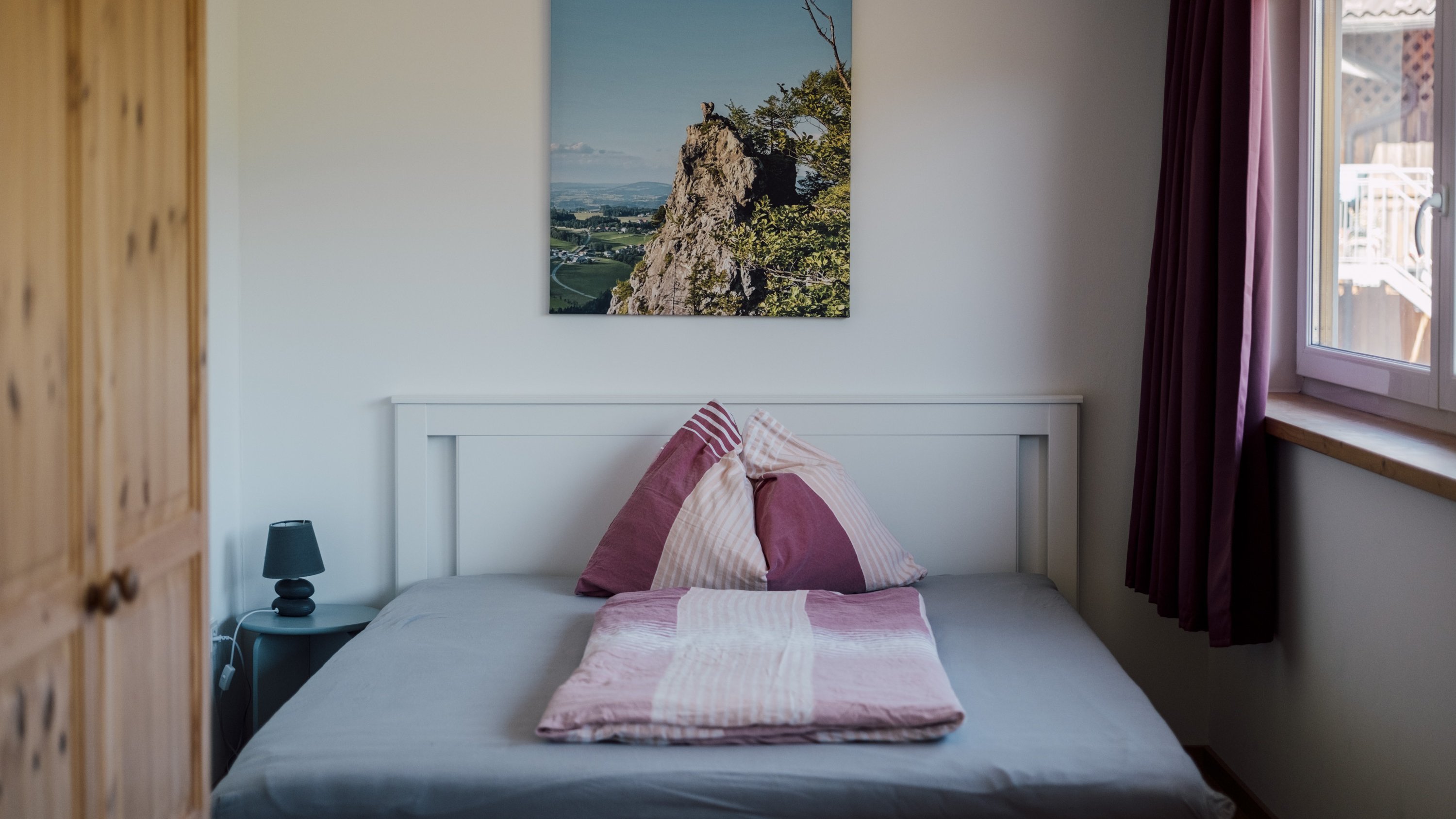 Ein Schlafzimmer mit einem Bett, das mit pink und grauer Bettwäsche dekoriert ist, daneben eine Lampe und ein Landschaftsgemä